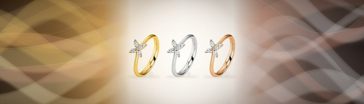 Ring Blume in 18 Karat Gelbgold, Weißgold und Roségold nebeneinander vor goldenen und roséfarbenen Seidenschleiern